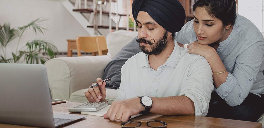 A man sitting down using a calculator and laptop, with a woman looking over his shoulder